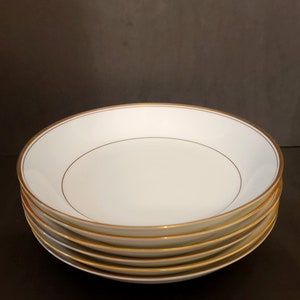 Può includere: Un set di quattro ciotole in ceramica bianca con bordo dorato. Le ciotole sono rotonde e hanno un leggero bordo.