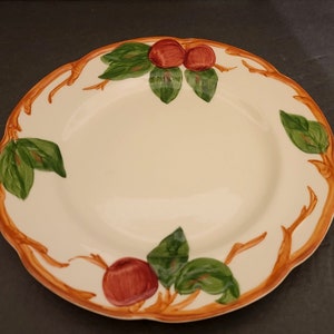 May include: A white ceramic dinner plate with a brown and orange rim. The plate features a design of green leaves and red apples on brown branches.