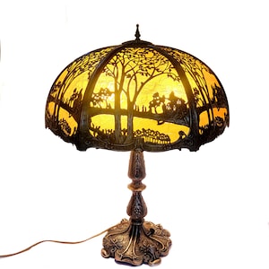 May include: A decorative table lamp with a bronze-colored base and a domed shade. The shade features a silhouette design of trees, buildings, and figures against a yellow background. The lamp has an ornate, detailed base and stem.