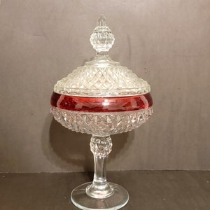 May include: A clear glass candy dish with a red band around the middle. The dish has a pedestal base and a domed lid with a finial.