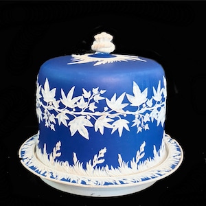 May include: A blue ceramic cake stand with a matching lid. The stand and lid are decorated with white leaf and floral patterns. The lid has a decorative handle. The stand sits on a white plate.