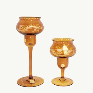 May include: Two amber-colored glass candle holders. The candle holders have a floral design in a lighter color. The taller candle holder has a long stem and a round base. The shorter candle holder has a shorter stem and a round base.