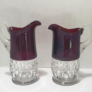May include: A set of two clear glass creamer pitchers with a deep red colored glass top. The pitchers have a diamond pattern on the bottom.