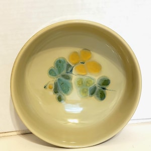 May include: A round, light yellow ceramic bowl with a floral design. The flowers are yellow and green, with a blue-green stem. The bowl has a simple, elegant design.