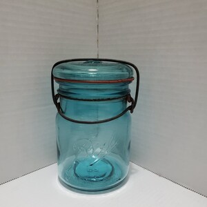 Op de afbeelding: Een blauwe glazen pot met een draadbeugelhandvat en een rood rubberen deksel. De pot heeft het woord "Ball" in reliëf aan de zijkant.