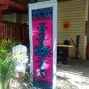 May include: A rectangular wall hanging with a pink center and white border. The pink section features a geometric design in black and turquoise. The design includes triangles, diamonds, and a central starburst pattern. The wall hanging is displayed outdoors.