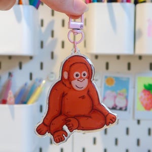 Puede incluir: Un llavero acrílico transparente con un orangután de dibujos animados en tonos naranja y marrón. El llavero se sostiene frente a una pared perforada blanca con varios útiles de arte y objetos decorativos.