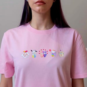 May include: A light pink t-shirt featuring embroidered cartoon characters in a row. The characters are in various colors, including white, blue, purple, and yellow. The shirt has short sleeves and a crew neck.