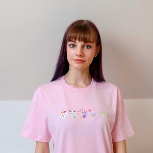 May include: A light pink t-shirt with a crew neck and short sleeves. The shirt features a row of colorful embroidered cartoon characters across the chest. The person in the image is wearing the shirt.