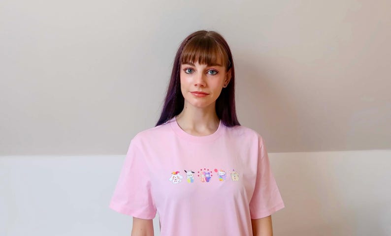 May include: A light pink t-shirt with a crew neck and short sleeves. The shirt features a row of colorful embroidered cartoon characters across the chest. The person in the image is wearing the shirt.
