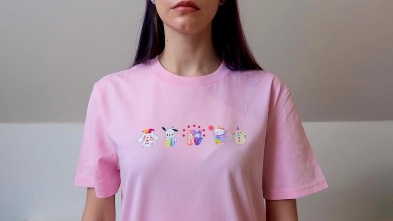 May include: A light pink t-shirt featuring embroidered cartoon characters in a row. The characters are in various colors, including white, blue, purple, and yellow. The shirt has short sleeves and a crew neck.