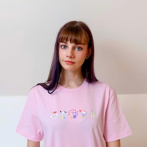 May include: A light pink t-shirt featuring an embroidered design of several cartoon characters. The shirt is made of a soft material and has short sleeves. The model is wearing the shirt.