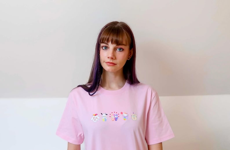 May include: A light pink t-shirt featuring an embroidered design of several cartoon characters. The shirt is made of a soft material and has short sleeves. The model is wearing the shirt.