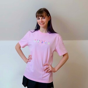 May include: A light pink t-shirt with a colorful floral design across the chest. The shirt is short-sleeved and appears to be made of a soft material. The text on the shirt is "ORCHIDS".