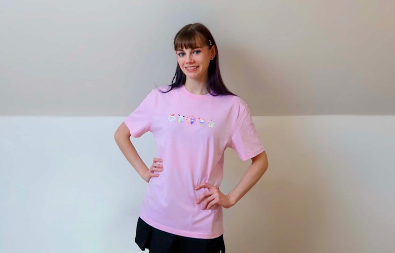 May include: A light pink t-shirt with a colorful floral design across the chest. The shirt is short-sleeved and appears to be made of a soft material. The text on the shirt is "ORCHIDS".