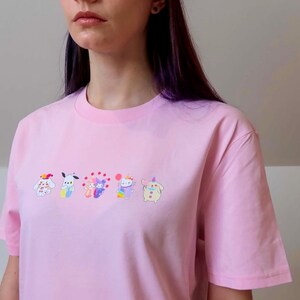 May include: A light pink t-shirt with a crew neck and short sleeves. The shirt features an embroidered design of six colorful cartoon characters across the chest. The characters have various colors and designs.