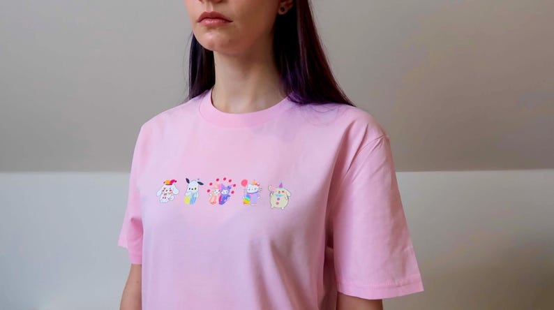 May include: A light pink t-shirt with a crew neck and short sleeves. The shirt features an embroidered design of six colorful cartoon characters across the chest. The characters have various colors and designs.