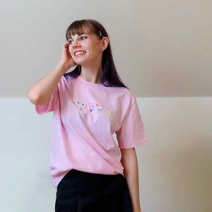 May include: A light pink t-shirt with a floral design. The shirt is short-sleeved and has a crew neck. The model is wearing black shorts.