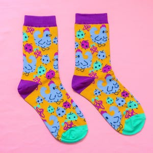 May include: A pair of patterned socks featuring light blue chicken illustrations, pink ladybugs, and teal star shapes on a yellow backdrop. The socks have purple cuffs, heels, and toes, with a teal trim.