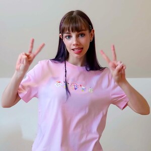 May include: A light pink t-shirt with a small, colorful embroidered design. The person is making a peace sign with both hands. The background is a plain, light-colored wall.