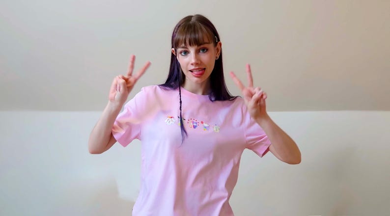 May include: A light pink t-shirt with a small, colorful embroidered design. The person is making a peace sign with both hands. The background is a plain, light-colored wall.
