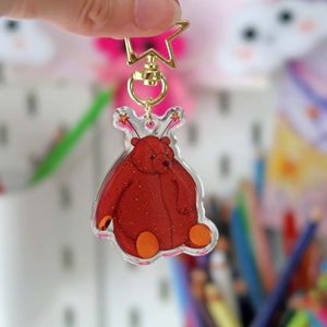 May include: A red glitter bear with orange feet and stars on its head is attached to a gold key chain. The bear is a clear acrylic charm.
