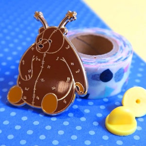 May include: A brown teddy bear pin with a stitched design and antenna-like appendages. The pin is on a blue background with white polka dots. The pin is accompanied by two yellow pin backs.