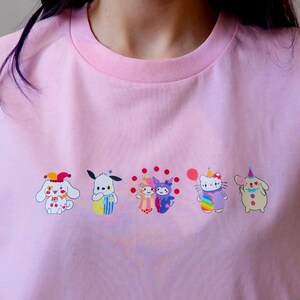 May include: Pink t-shirt featuring a row of colorful cartoon characters. The characters include a white dog with a clown hat, a blue and yellow dog, two figures with red polka dots, a Hello Kitty character with a rainbow outfit, and a beige character with a party hat.