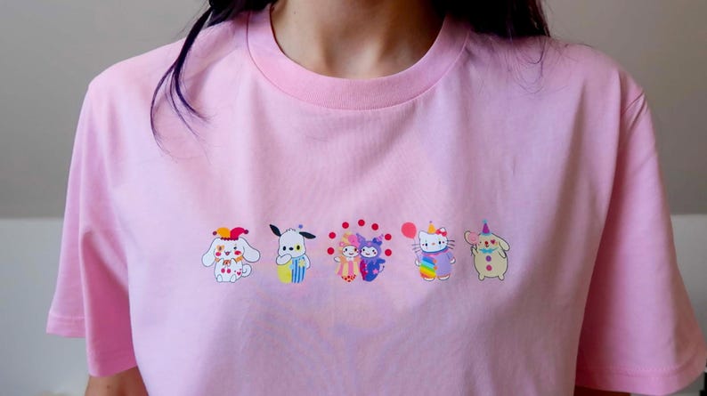 May include: Pink t-shirt featuring a row of colorful cartoon characters. The characters include a white dog with a clown hat, a blue and yellow dog, two figures with red polka dots, a Hello Kitty character with a rainbow outfit, and a beige character with a party hat.