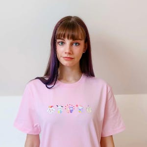 May include: A light pink t-shirt featuring a row of colorful cartoon characters embroidered across the chest. The characters include a dog, a bunny, a bear, a cat, and a cupcake. The shirt has short sleeves and a crew neck.