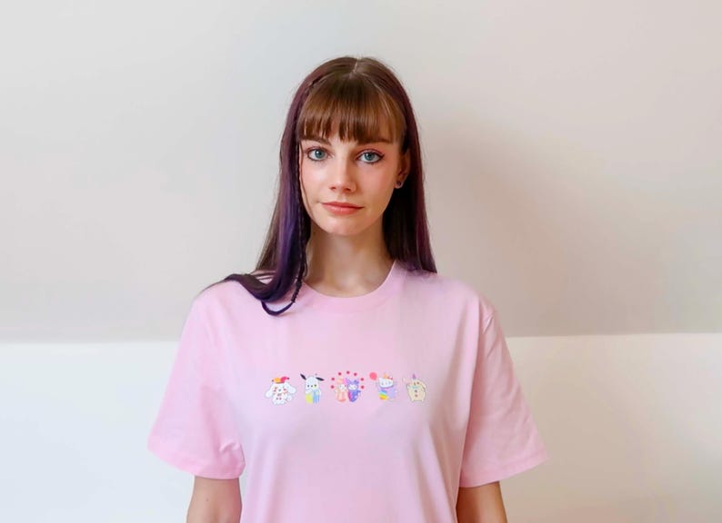 May include: A light pink t-shirt featuring a row of colorful cartoon characters embroidered across the chest. The characters include a dog, a bunny, a bear, a cat, and a cupcake. The shirt has short sleeves and a crew neck.