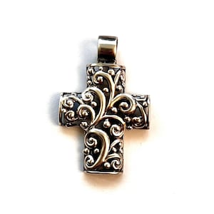 May include: A silver-colored pendant in the shape of a cross. The cross features an ornate, swirling design with a dark background. The bail is a simple, rectangular shape. The pendant is likely made of metal and is displayed against a white background.