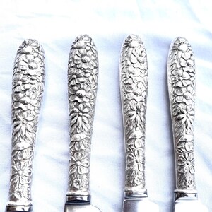May include: Four silver butter knives with intricately carved floral designs on the handles. The handles are covered in detailed relief patterns of various flowers and leaves. The blades are smooth and shiny, reflecting the light.