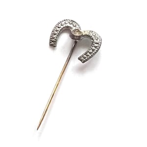 May include: A vintage silver-toned horseshoe pin with a long, gold-colored pin. The horseshoe design is detailed with small, raised dots. The pin is likely an antique or vintage accessory.