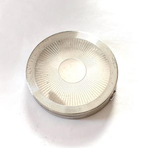 May include: A round, silver-toned metal compact case featuring a sunburst pattern of fine, radiating lines etched into the surface. The center of the lid has a smooth, circular polished area, and the outer edge shows signs of wear and light scratches.