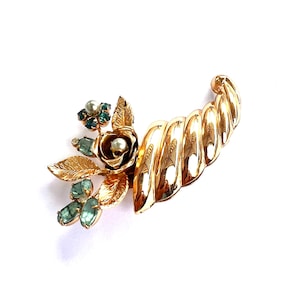 May include: A gold-toned vintage brooch featuring a floral design with a rose, leaves, and teal-colored gemstones. The brooch has a curved, textured element and a pearl accent. A unique piece of jewelry.