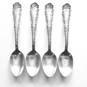 May include: Four silver spoons with ornate, scrolling handles and oval bowls are arranged on a white background. The spoons have a reflective surface, and the handles feature a decorative design. The spoons are likely for serving or eating.