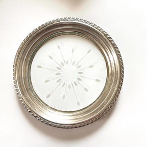 May include: A round coaster featuring a metallic silver rim with a decorative ridged edge and a circular white glass center. The glass insert displays a frosted starburst pattern radiating from the middle, consisting of thin lines and leaf-shaped tips.