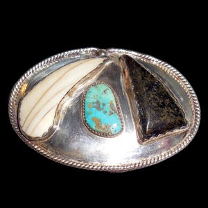 May include: Oval silver belt buckle with three inlaid stones. Features a turquoise stone, a white stone, and a black stone. The buckle has a rope-like border and is set against a black background.