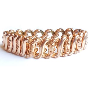 May include: A gold-toned, vintage bracelet with an ornate, repeating link design. The links are curved and feature detailed scrollwork, creating a textured and elegant appearance. The bracelet is shown against a white background.