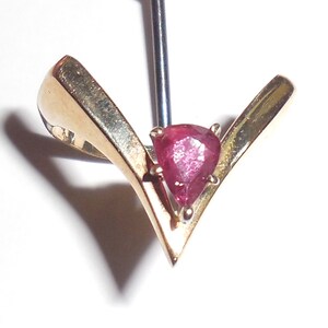 May include: A gold-toned pendant with a pear-shaped, deep red gemstone at its center. The pendant has a V-shaped design, with the gemstone held in place by small prongs. The metal has a polished finish.