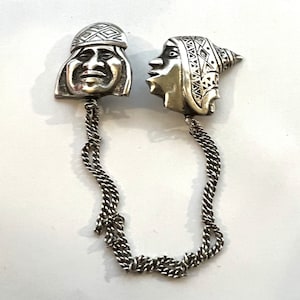 May include: A pair of silver-toned pendants featuring stylized human faces with intricate geometric patterns. The pendants are connected by a silver chain.