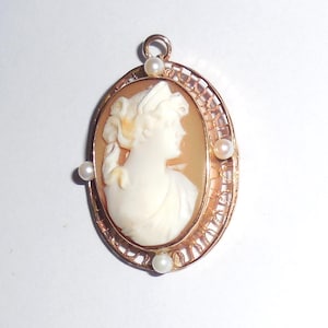 May include: Oval cameo pendant with a profile of a woman in white against a tan background. The cameo is set in a gold filigree frame and accented with three small pearls. A small loop is at the top for hanging.