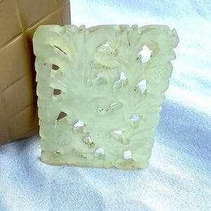 May include: A rectangular, pale green jade carving with a floral design. The carving features intricate details of leaves and flowers, with negative space creating an openwork effect. The jade has a smooth, polished finish.