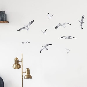 Puede incluir: Un interior de sala de estar con un sof&aacute; azul, una mesa de caf&eacute; de madera, una l&aacute;mpara de pie, una c&oacute;moda y una planta en maceta. La pared est&aacute; decorada con una bandada de calcoman&iacute;as de gaviotas blancas.