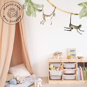 Può includere: Un muro bianco con due scimmie appese a una vite con foglie verdi. Le scimmie sono marroni e grigie. C'è una tenda a baldacchino marrone chiaro nell'angolo in basso a sinistra dell'immagine. Uno scaffale in legno con contenitori bianchi e libri si trova nell'angolo in basso a destra dell'immagine. Il testo "Plastic-Free Wall Stickers" è in un cerchio nell'angolo in alto a sinistra dell'immagine.