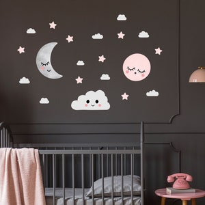 May include: A gray crib with a pink blanket and a gray pillow. The wall behind the crib has pink and white clouds, stars, and moons with smiling faces.