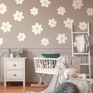 Peut inclure: Un berceau blanc avec une couverture blanche et un ours en peluche sur le dessus. Le berceau est dans une chambre d'enfant avec un mur gris et des autocollants de marguerites blanches. Il y a une table de chevet blanche à deux tiroirs à côté du berceau.