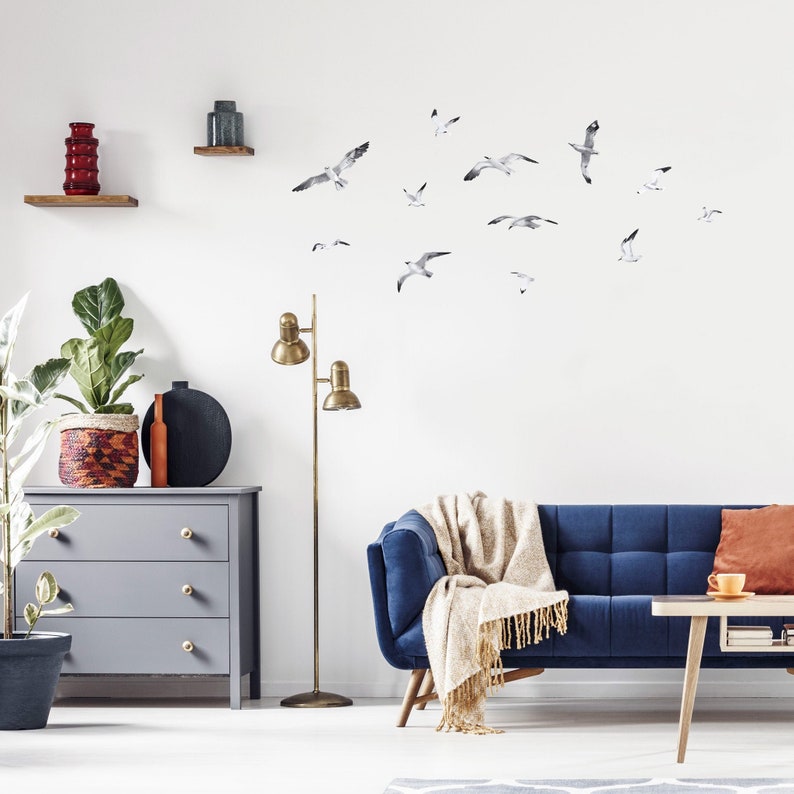 Puede incluir: Un interior de sala de estar con un sof&aacute; azul, una mesa de caf&eacute; de madera, una l&aacute;mpara de pie, una c&oacute;moda y una planta en maceta. La pared est&aacute; decorada con una bandada de calcoman&iacute;as de gaviotas blancas.