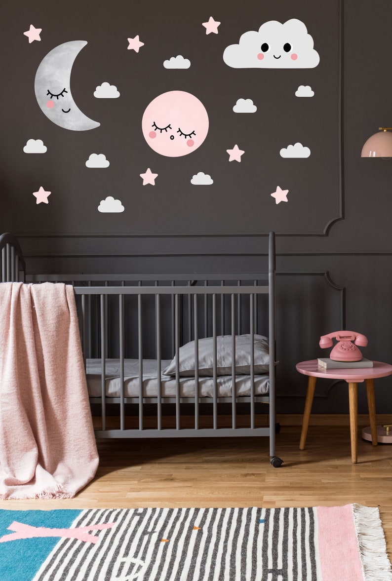 May include: A gray crib with a pink blanket and a gray pillow. The wall behind the crib is dark gray and has pink and white clouds, stars, and a moon and sun with faces on them.